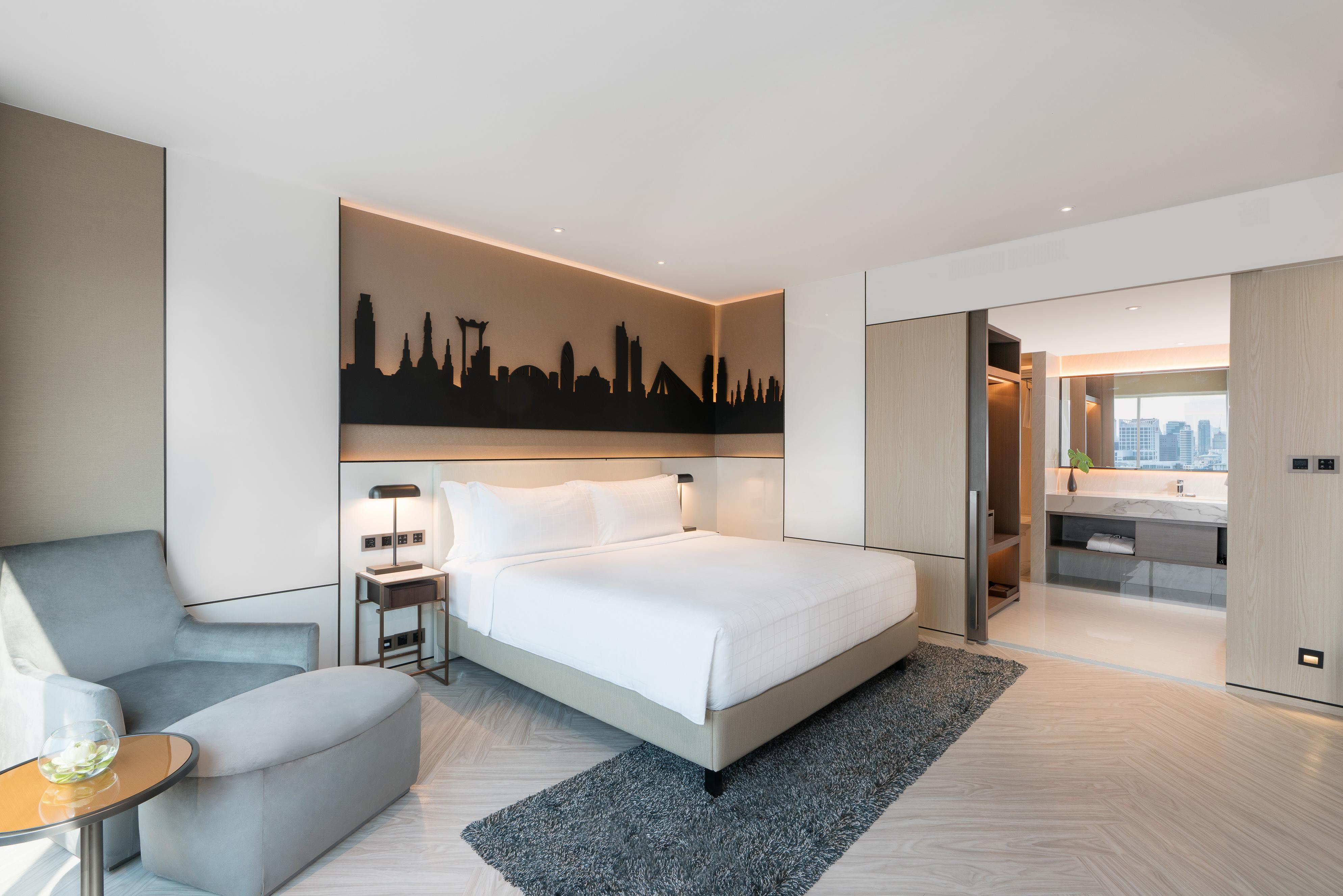 Pathumwan Princess Hotel - Sha Extra Plus Certified Bangkok Exterior foto The photo shows a modern hotel room featuring a minimalistic design. There is a large, white bed with a headboard that has a silhouette of a city skyline illuminated by soft lighting. To the left, there is a comfortable gray armchair next to a small 