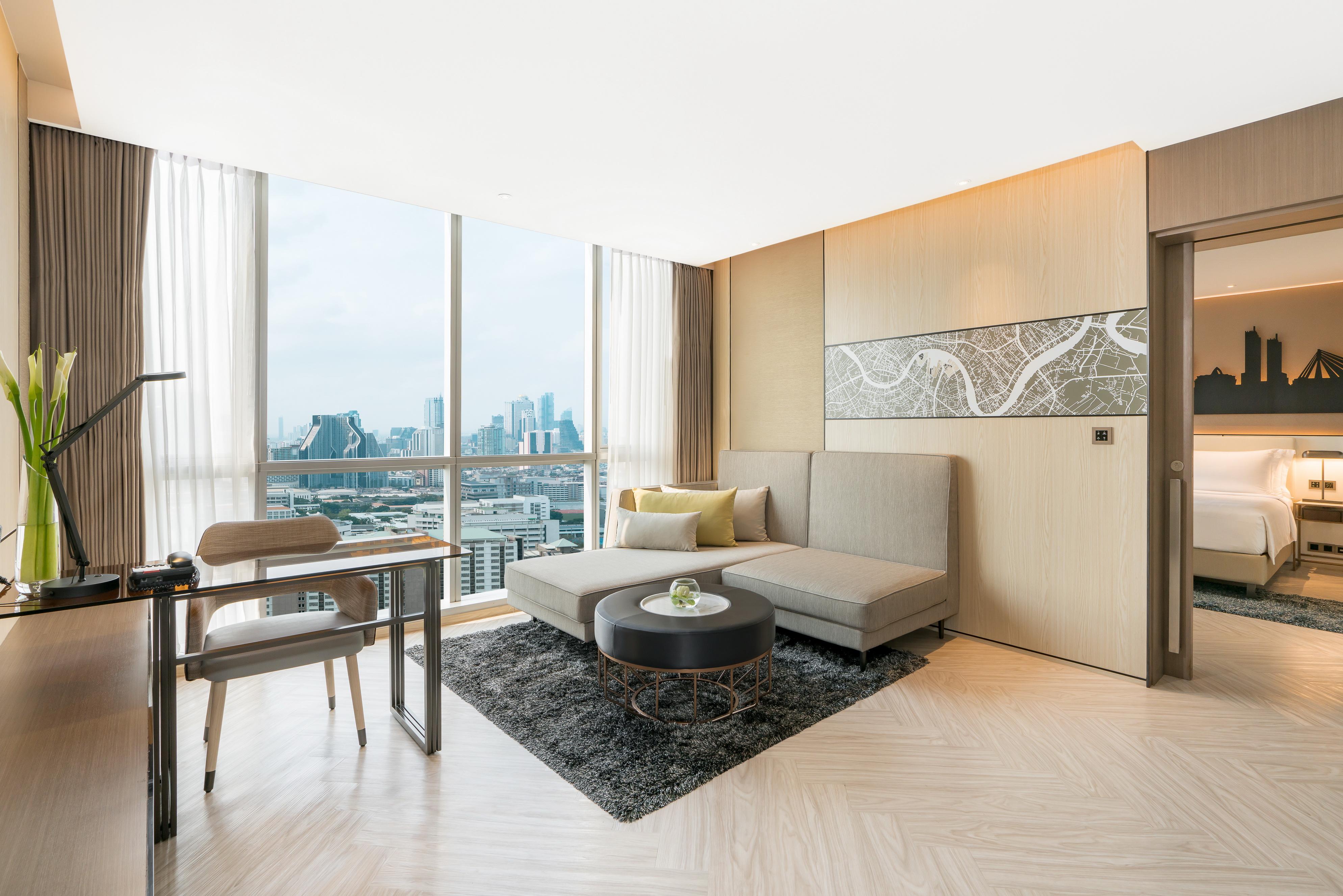 Pathumwan Princess Hotel - Sha Extra Plus Certified Bangkok Exterior foto The photo shows a modern, stylish living area in a hotel or apartment. It features a large window that offers a view of a city skyline. The room is designed with neutral-colored walls and light wooden flooring. There is a comfortable gray sofa with t