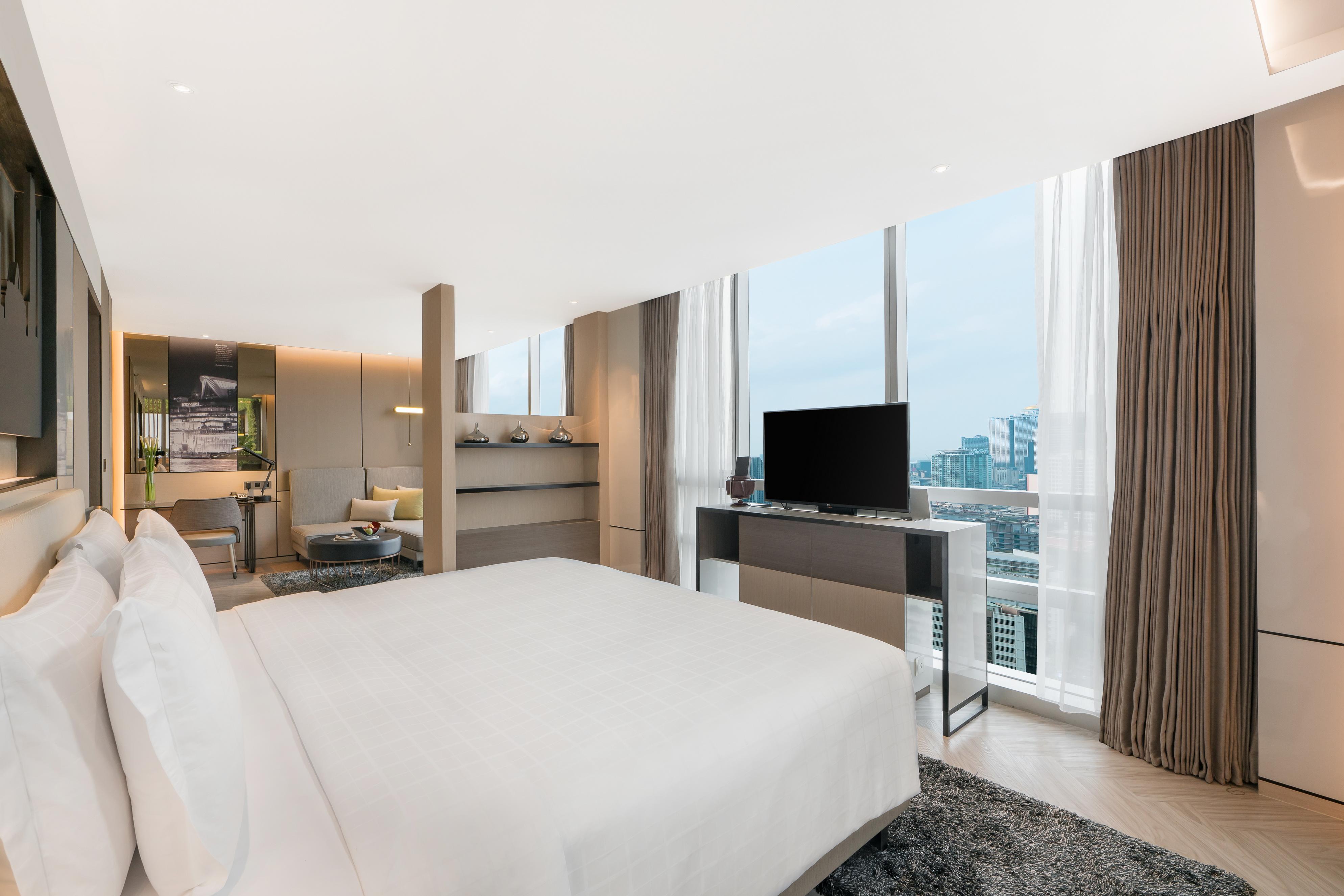 Pathumwan Princess Hotel - Sha Extra Plus Certified Bangkok Exterior foto The photo shows a modern hotel room, featuring large windows that provide a view of the city. The room has a spacious layout with a king-sized bed covered in white linens, a bedside table, and a contemporary television mounted on a console. There are
