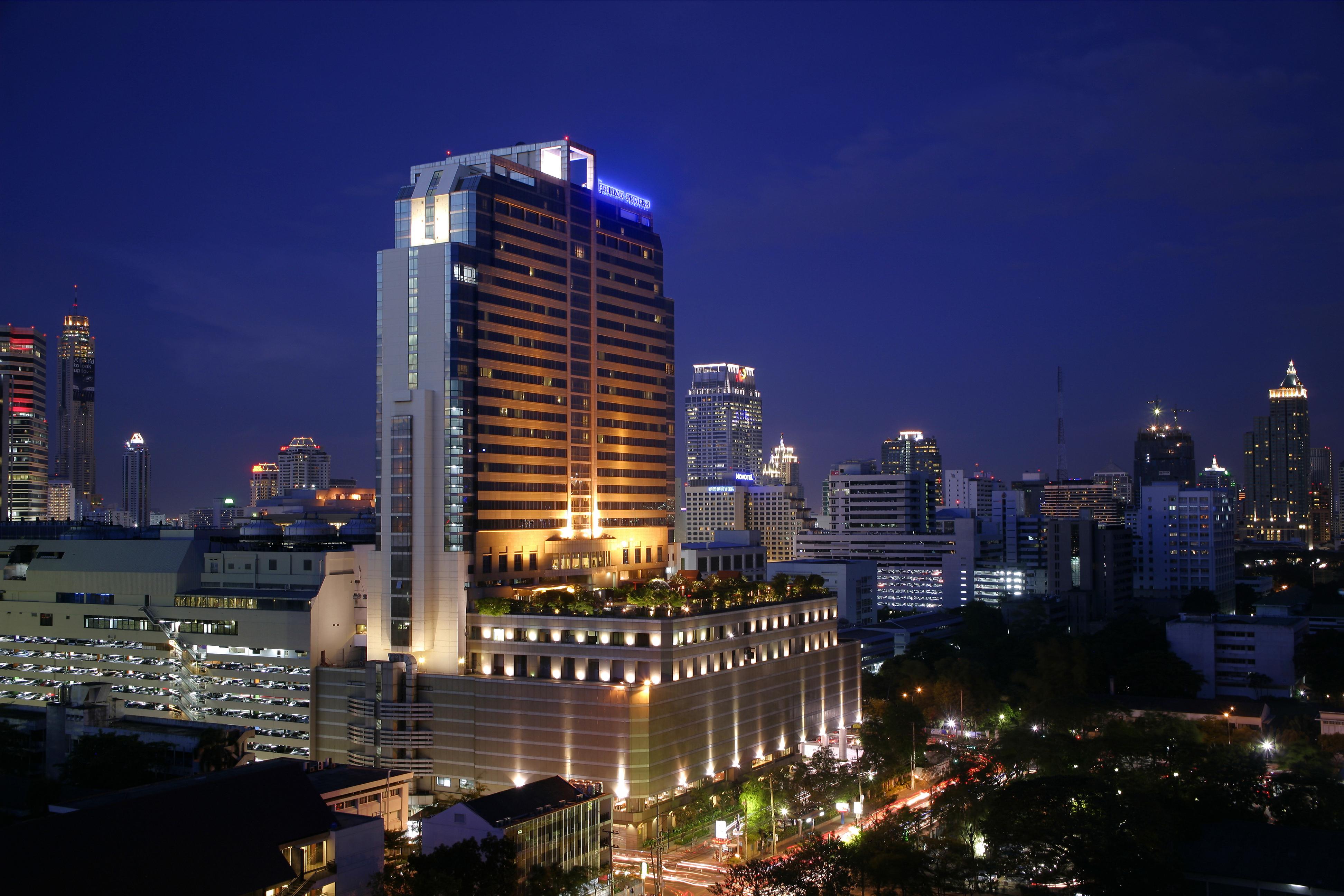 Pathumwan Princess Hotel - Sha Extra Plus Certified Bangkok Exterior foto The photo shows a cityscape at night, featuring a tall, modern hotel building illuminated with bright lights. The building has a sleek design and is surrounded by a variety of other skyscrapers and urban structures. The street below is bustling with 