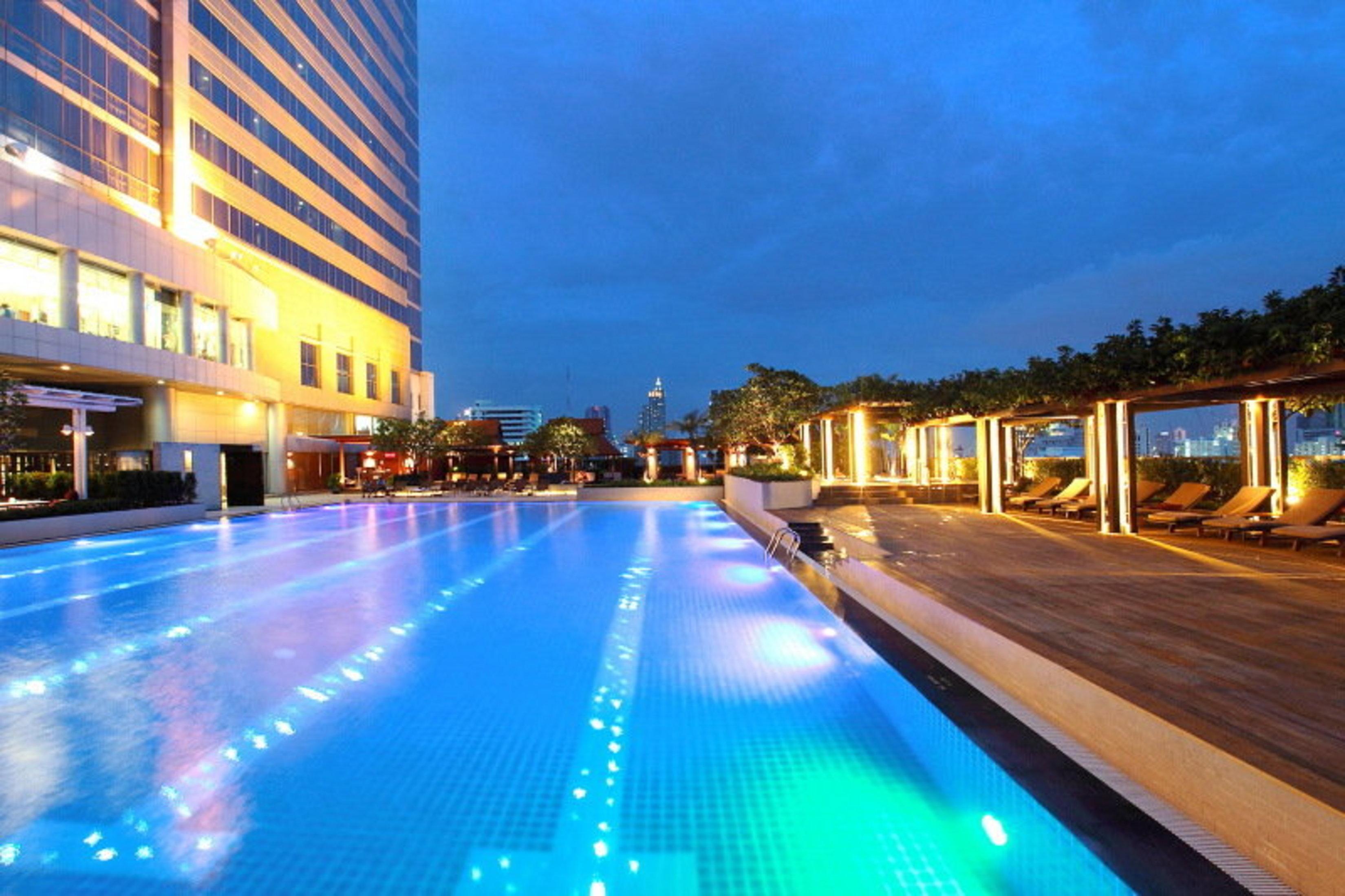 Pathumwan Princess Hotel - Sha Extra Plus Certified Bangkok Exterior foto The photo showcases a luxurious swimming pool at dusk. The pool is illuminated with colorful lights that create a vibrant atmosphere. Surrounding the pool are lounge areas with wooden decking and lounge chairs, providing a relaxing space for guests. 