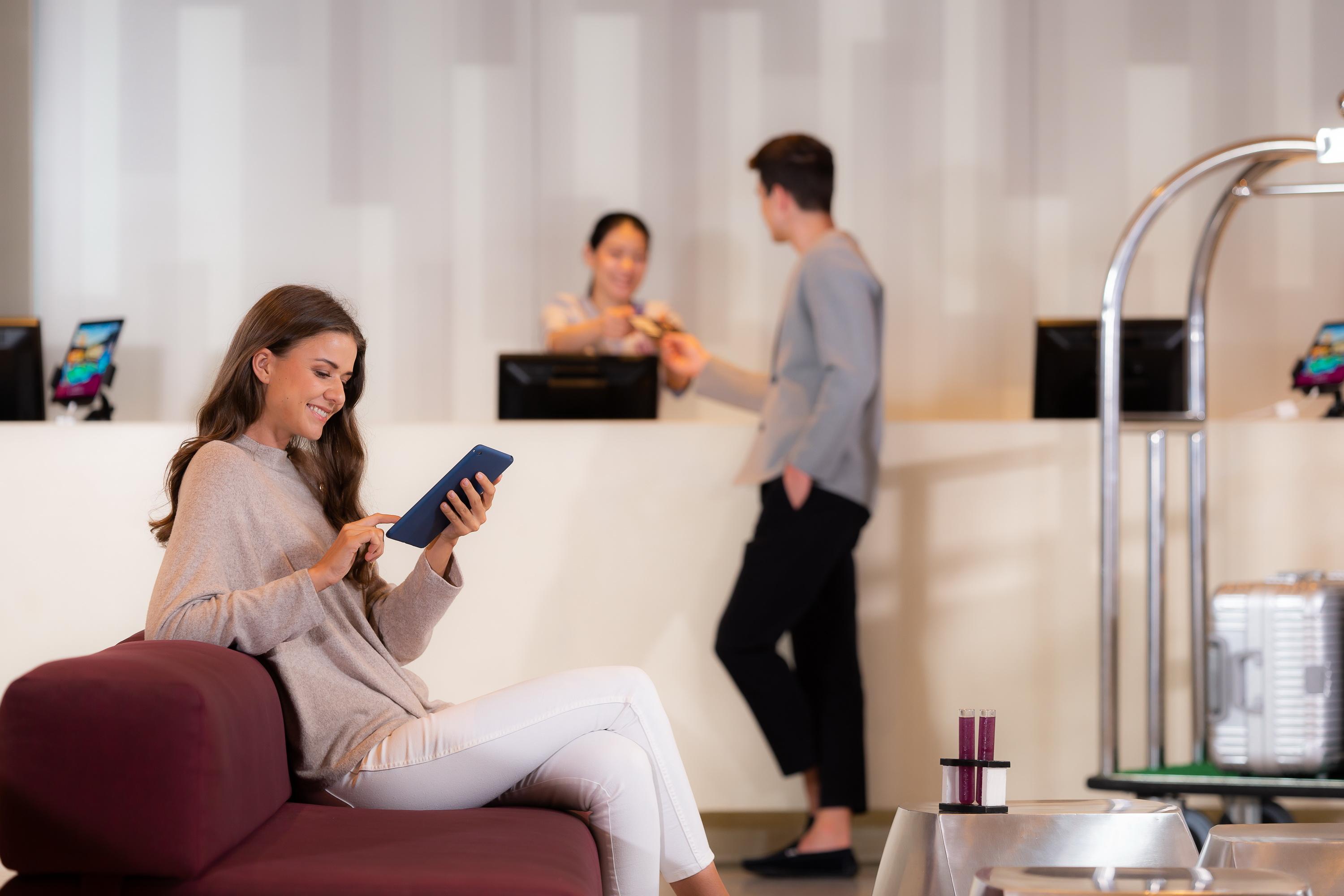 Pathumwan Princess Hotel - Sha Extra Plus Certified Bangkok Exterior foto The photo shows a modern reception area. In the foreground, a woman is sitting on a maroon sofa, engaged with a tablet. She is wearing a light sweater and white pants. In the background, two people are at the reception desk. One person is a staff mem