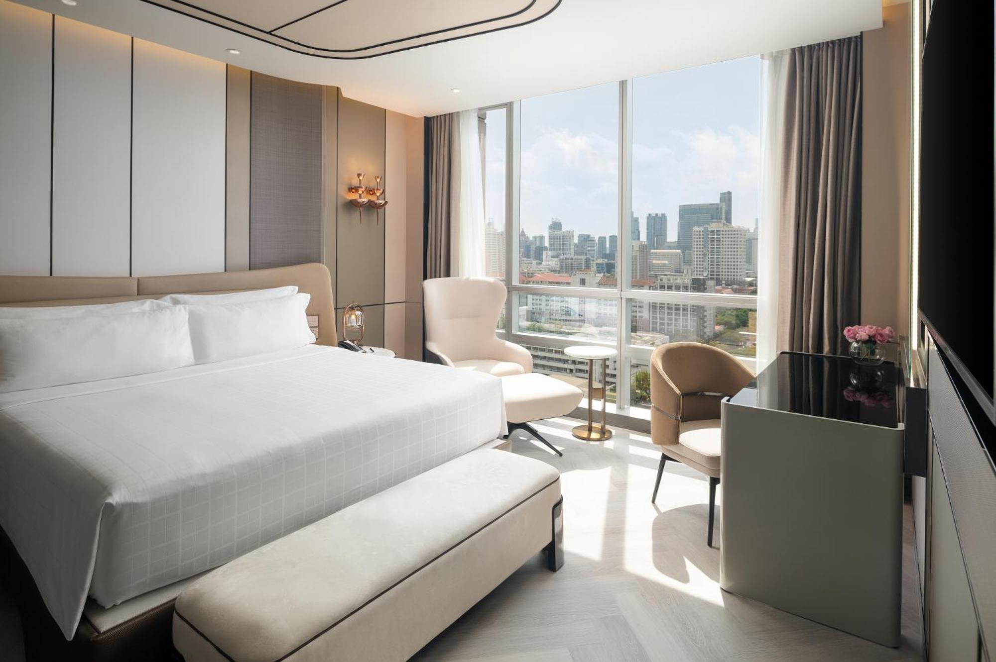 Pathumwan Princess Hotel - Sha Extra Plus Certified Bangkok Exterior foto The photo shows a modern hotel room featuring a large bed with white linens. There are two stylish chairs and a small table positioned near a large window that offers a view of the city skyline. A sleek desk is also present, with flowers placed on it