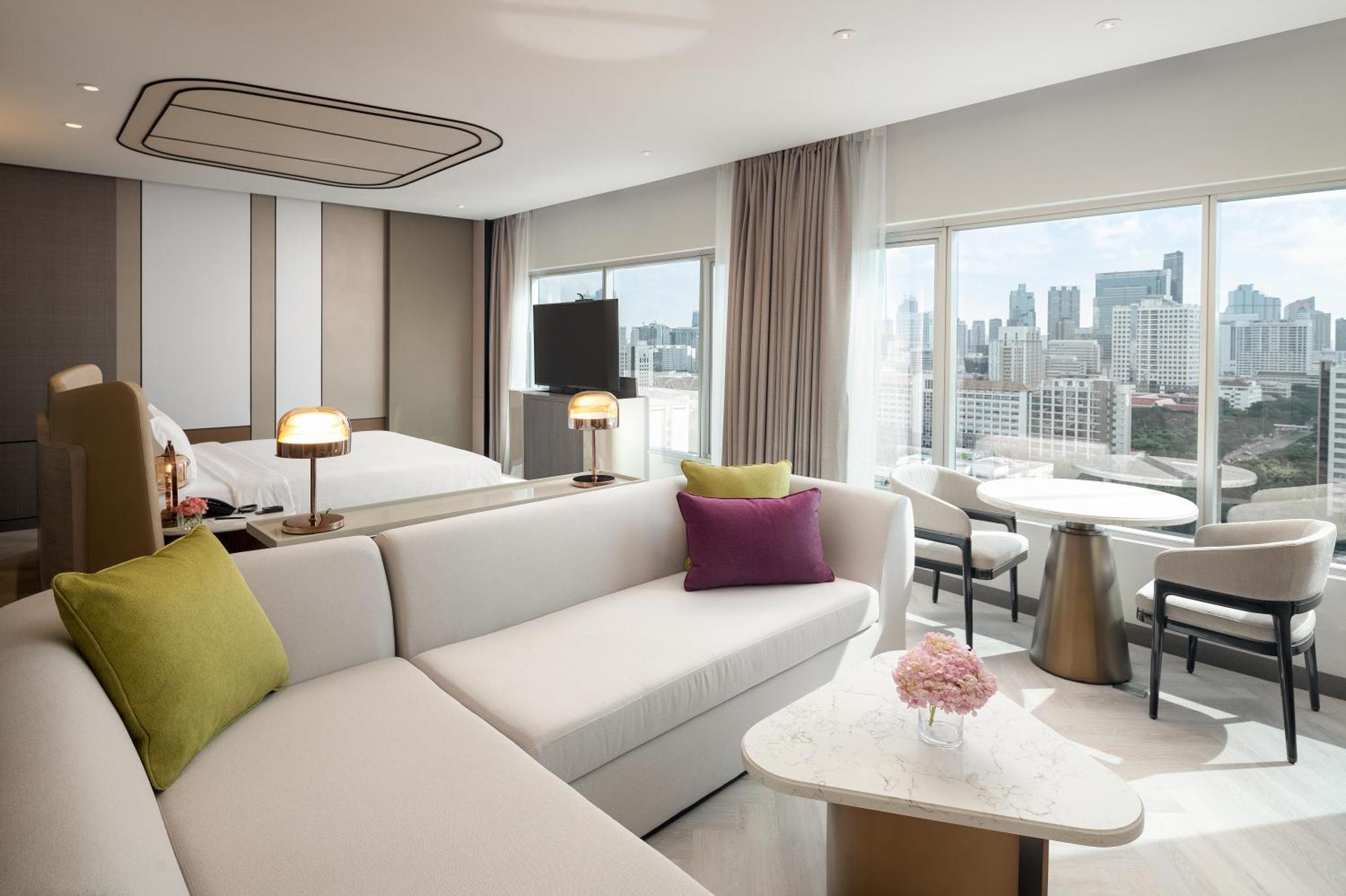 Pathumwan Princess Hotel - Sha Extra Plus Certified Bangkok Exterior foto The photo showcases a modern hotel room featuring a stylish interior design. There is a large, comfortable seating area with a white sofa adorned with colorful cushions in green and purple. Nearby, a small round table with a floral centerpiece stands