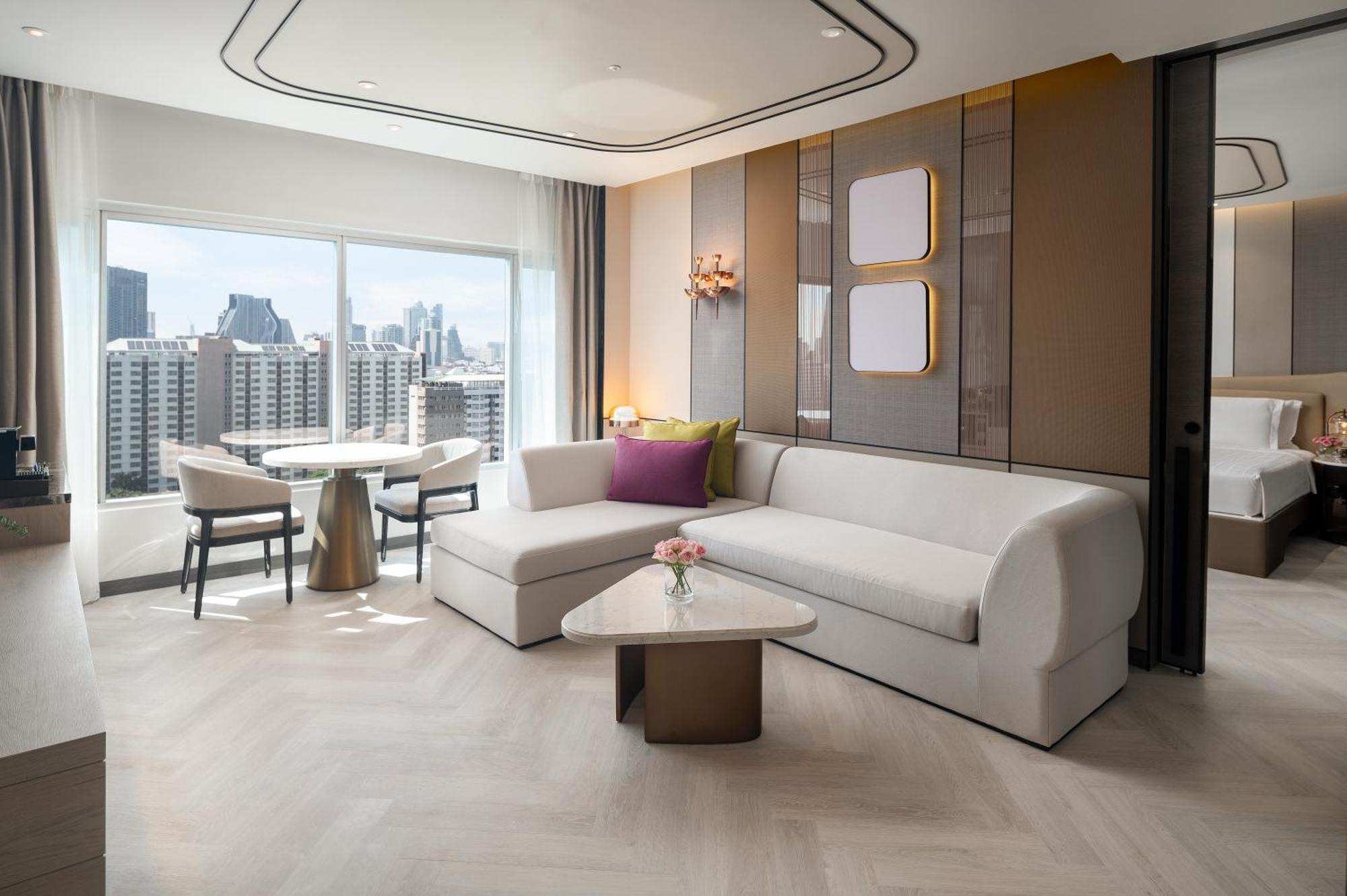 Pathumwan Princess Hotel - Sha Extra Plus Certified Bangkok Exterior foto The photo shows a modern and stylish hotel room or living space. There is a comfortable white sofa with some decorative cushions, and a small coffee table in front of it. A round dining table with chairs is visible nearby. Large windows provide a vie