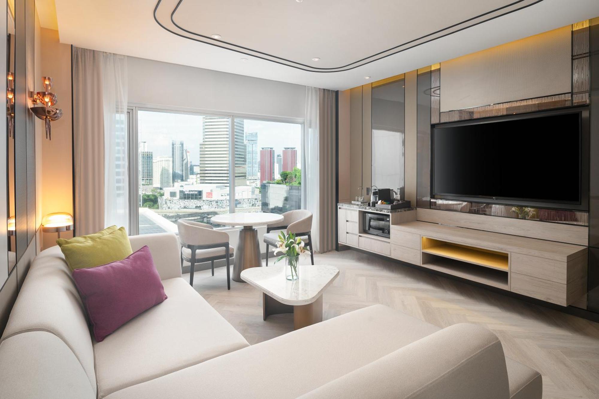 Pathumwan Princess Hotel - Sha Extra Plus Certified Bangkok Exterior foto The photo shows a modern and stylish hotel room or living area. There's a light-colored sofa with decorative pillows in shades of purple and beige. In front of the sofa, there's a small coffee table. To one side, there is a dining table with chairs, 