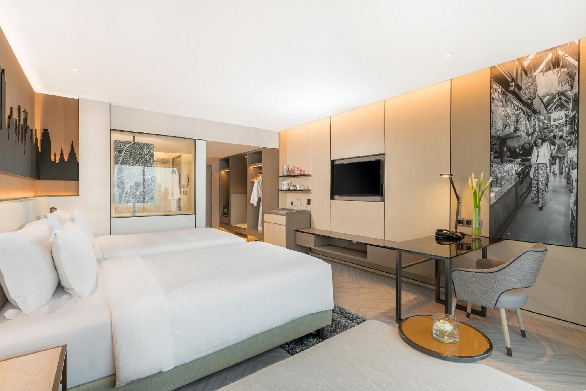 Pathumwan Princess Hotel - Sha Extra Plus Certified Bangkok Zimmer foto The photo shows a modern hotel room featuring a minimalist design. There are two beds with white bedding, positioned against one wall. A stylish desk and chair are situated near a large window, which allows natural light to fill the space. A flat-scr