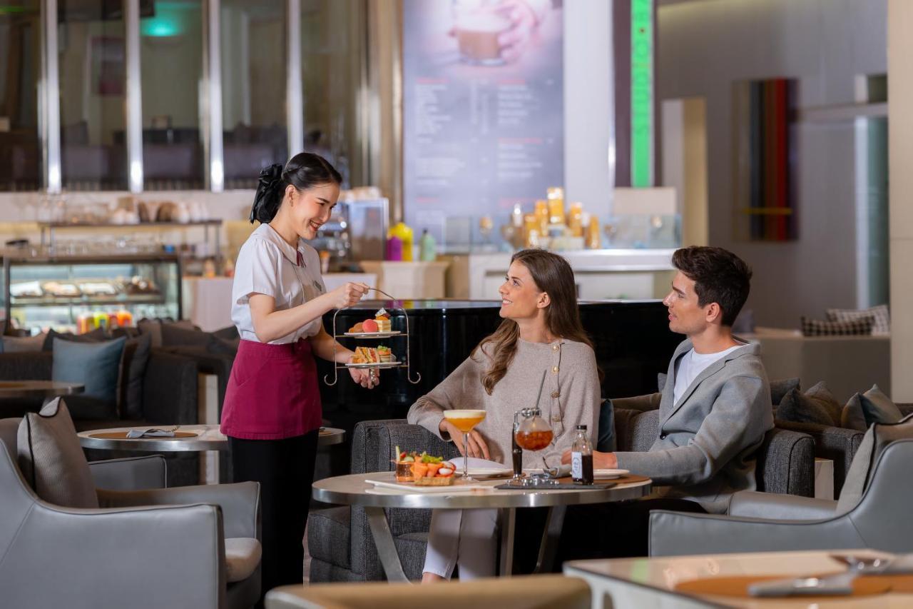 Pathumwan Princess Hotel - Sha Extra Plus Certified Bangkok Exterior foto The photo shows a scene in a stylish cafe or restaurant. A waitress is serving a dessert stand to two guests who are seated at a table. The waitress is dressed in a uniform and appears to be presenting a selection of desserts. The two guests, a man a